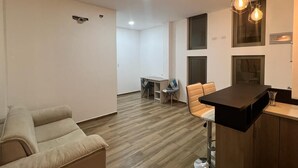 Een moderne woonkamer met een bank, een eethoek met een tafel en stoelen, en een keuken met een aanrecht en hangende verlichtingsarmaturen.