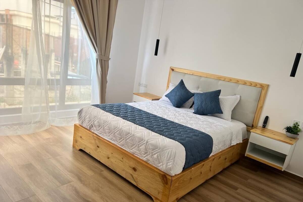 Una cama de madera con colcha blanca y azul, una mesita de noche con una planta, y una ventana con cortinas de tul.