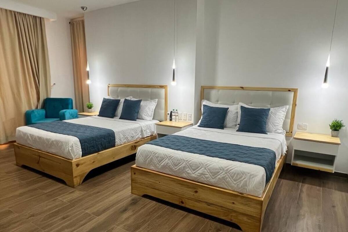Dos camas dobles con cabeceros de madera y ropa de cama blanca, un sillón color turquesa y una mesita de noche con lámpara.