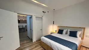 Een slaapkamer met een houten bedframe, een witte deur en een gang die leidt naar een keuken.