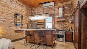 A kitchen with a brick wall, wooden bar stools, a stainless steel refrigerator, and a large island with a hanging light fixture.