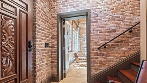 A hallway with a wooden door and a brick wall, leading to a staircase with wooden steps and a metal handrail.