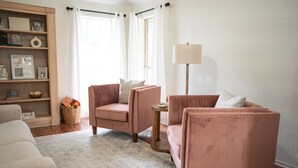 A living room with a beige sofa, a pink armchair, a small round table, and a floor lamp. There is a bookshelf with framed photos and a basket with books. - A Cozy Sooner Stay (Norman)