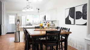 A spacious kitchen with white cabinets, a wooden dining table, and black chairs. There are two pendant lights above the table and a trash can near the door. - A Cozy Sooner Stay (Norman)