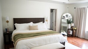 A neatly arranged bedroom with a large bed, bedside tables, a bench, a mirror, and a door leading to another room. - A Cozy Sooner Stay (Norman)