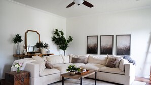 A living room with a sectional sofa, a coffee table, a wooden side table, a mirror, and three abstract paintings on the wall. - A Cozy Sooner Stay (Norman)