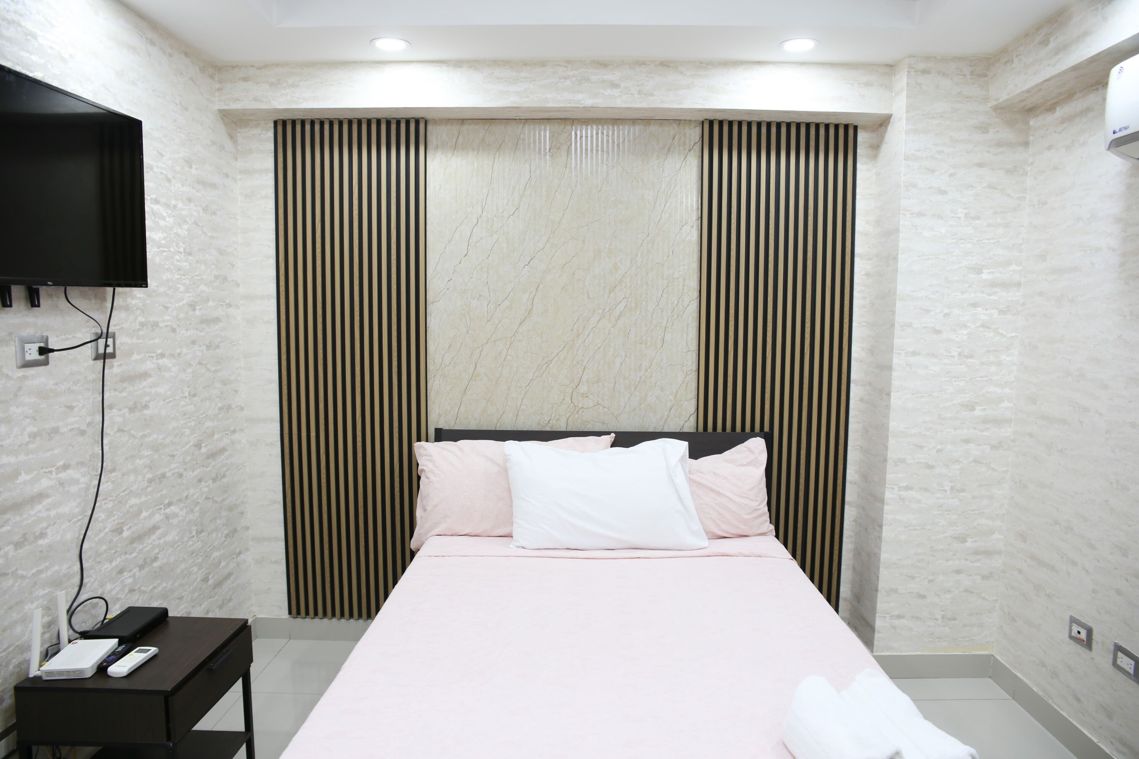 Quarto com cama, televisão fixada na parede e mesa de cabeceira com abajur e telefone.