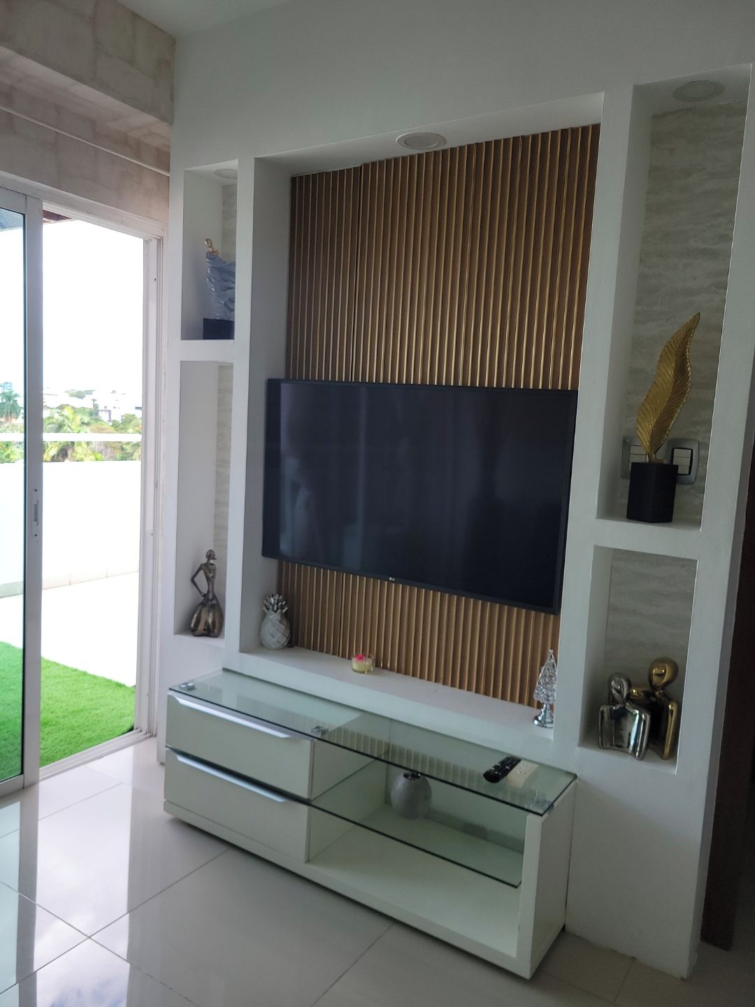 A modern living room with a glass TV stand, a flat-screen TV mounted on a wall with vertical blinds, and a glass cabinet below.