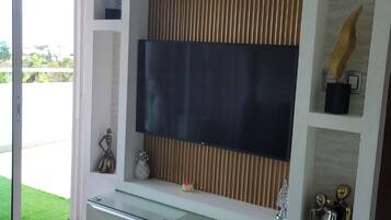 A modern living room with a glass TV stand, a flat-screen TV mounted on a wall with vertical blinds, and a glass cabinet below.