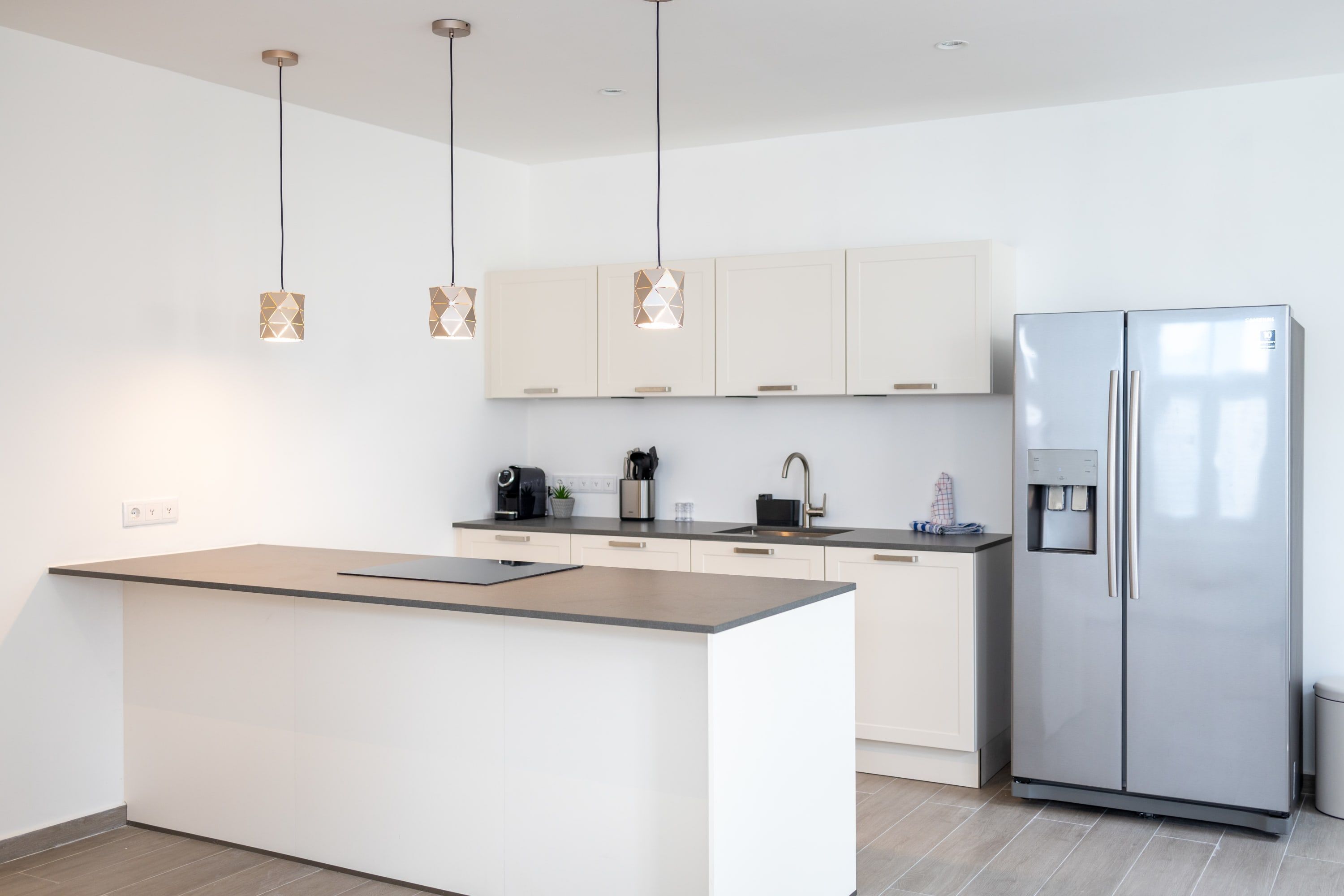 A modern kitchen with a central island, stainless steel refrigerator, pendant lights, and light-colored cabinets.