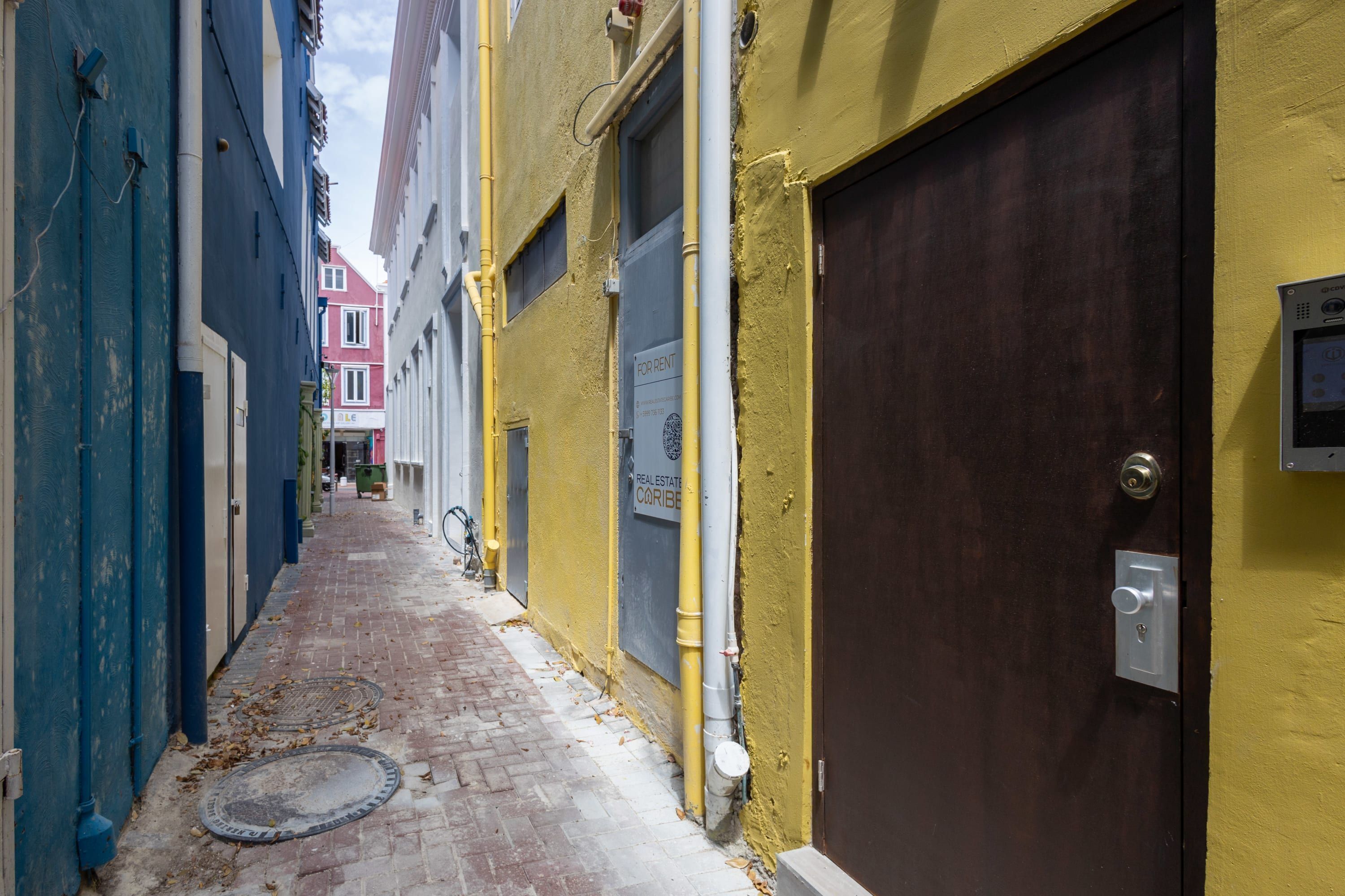 Un callejón estrecho flanqueado por edificios, una puerta a la vista con cerradura y una tapa de alcantarilla en el suelo.