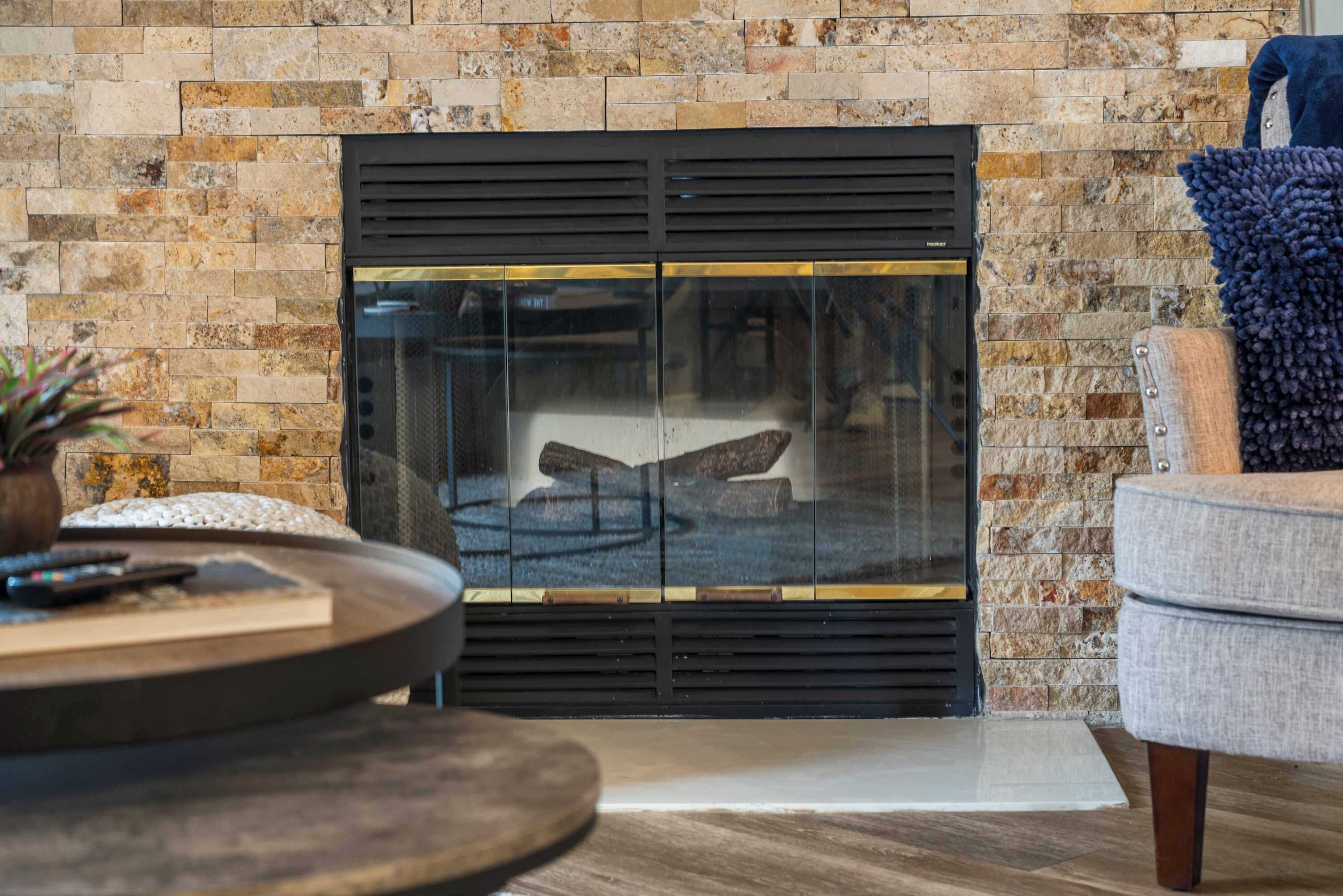 A fireplace with a glass door, a round wooden table with a remote control, and a grey armchair.