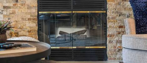 A fireplace with a glass door, a round wooden table with a remote control, and a grey armchair.