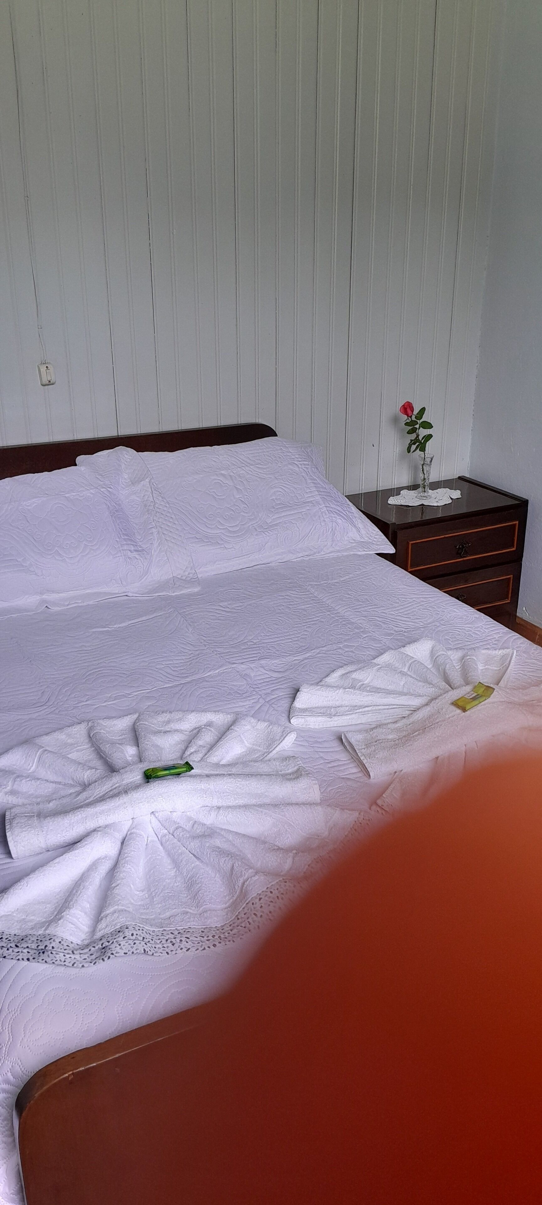 A neatly made bed with a white bedspread, a small vase with a single red rose on a nightstand, and a wooden dresser with drawers.