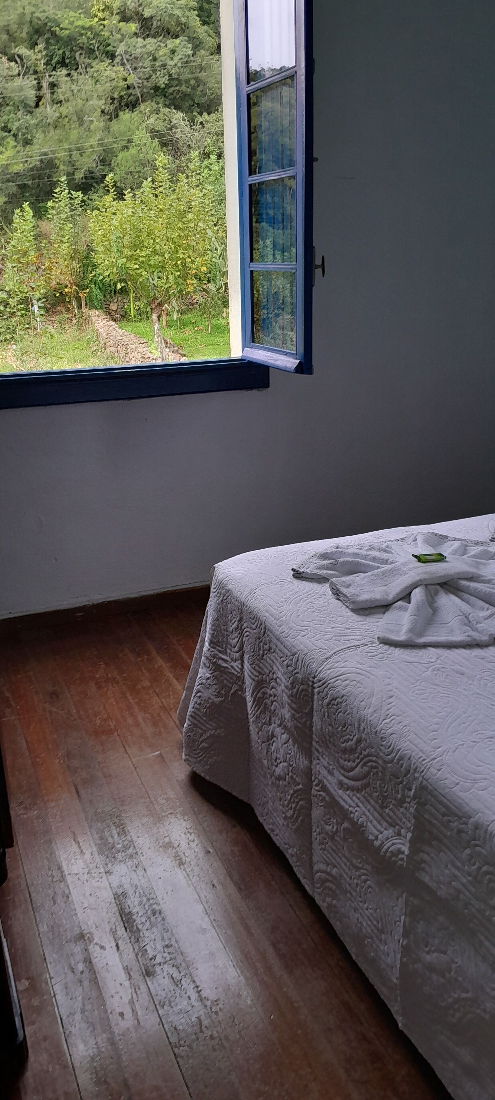 Una habitación con piso de madera, una cama con cubierta blanca y una ventana con vista a follaje.