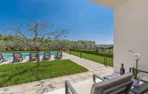 A patio with a pool, lounge chairs, a small table with a plant and wine glasses, and a view of the pool and greenery.