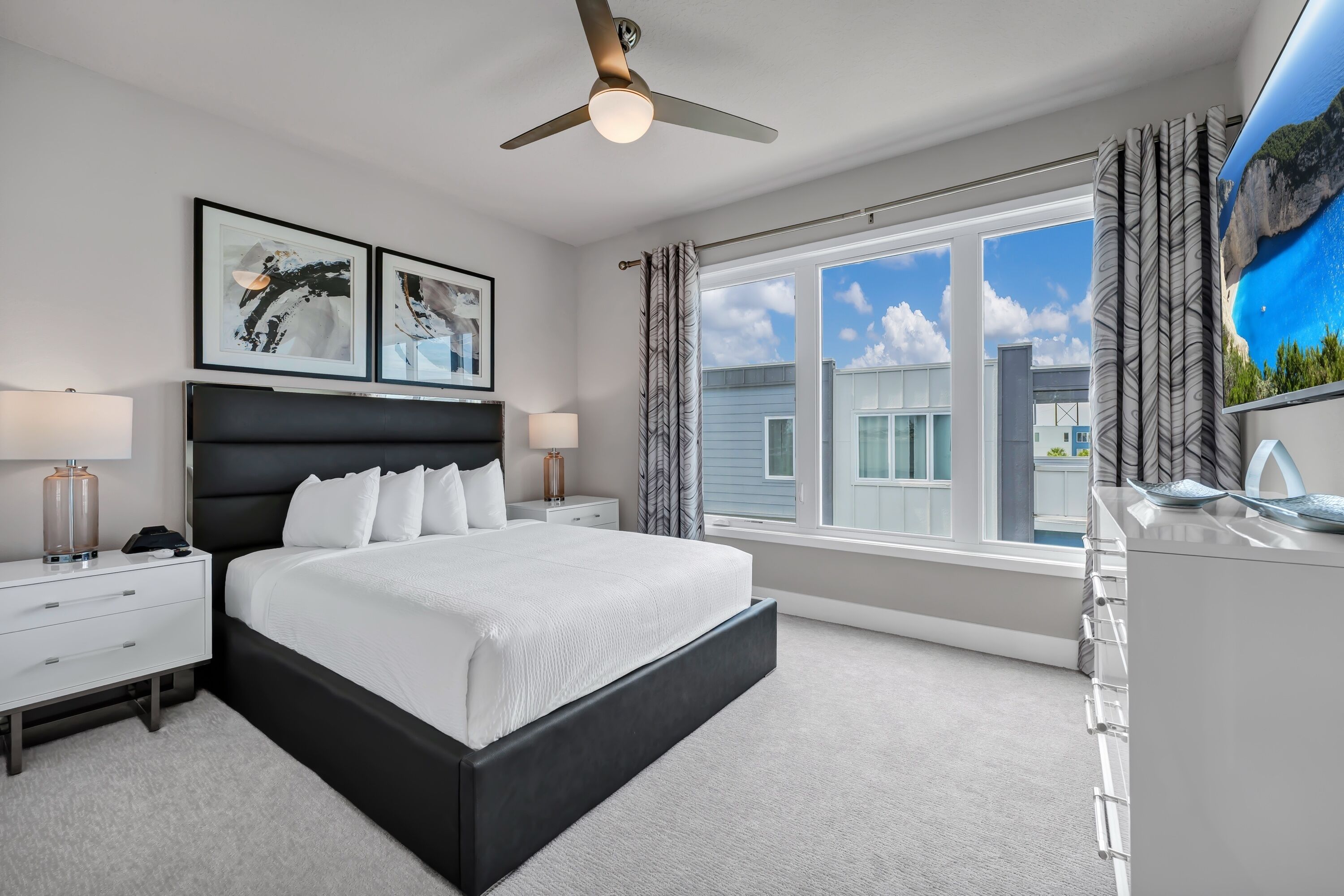A modern bedroom with a large bed, a ceiling fan, a window with curtains, and artwork on the wall.