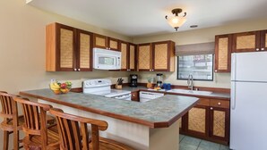 A kitchen with wooden cabinets, a white refrigerator, a microwave, a stove, and a sink. There is a fruit bowl on the counter and a window above the sink.