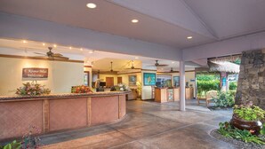 A reception area with a curved wooden counter, a ceiling fan, and a sign that reads "E Komo Mai Welcome".
