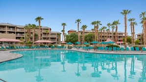 A hotel pool area with multiple lounge chairs and umbrellas, surrounded by palm trees and a multi-story building. - Desert Oasis: Spacious 2-Bedroom Suite at Club Wyndham Indio (Indio)