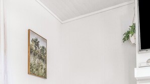 A room with a wooden floor, a grey upholstered armchair, a white vase with a plant, a woven pouf, a framed picture on the wall, and a white cabinet with a plant on top. - The P'Cola Sunrise. 1 bed 1 bath cottage (Pensacola)