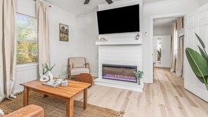 A living room with a fireplace, a television mounted above it, a ceiling fan, and a view into another room through an open door. - The P'Cola Sunrise. 1 bed 1 bath cottage (Pensacola)