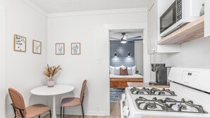 A compact kitchen with a gas stove, a microwave, and a coffee maker. There is a dining area with two chairs and a small table. The walls are white, and there are framed pictures on the wall. - The P'Cola Sunrise. 1 bed 1 bath cottage (Pensacola)