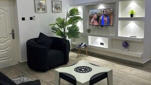 Una sala de estar moderna con un sofá negro, una mesa de centro blanca, un televisor montado en la pared y una planta decorativa.