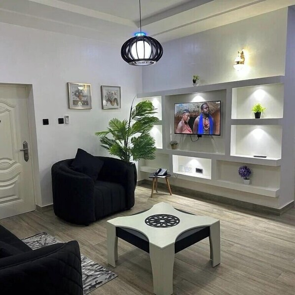 A modern living room with a black sofa, a white coffee table, a TV mounted on the wall, and a decorative plant.