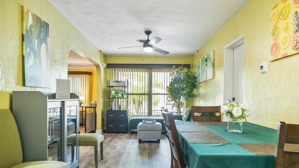 Un coin repas avec une table dressée pour deux personnes, un ventilateur de plafond et une grande fenêtre avec des rideaux.