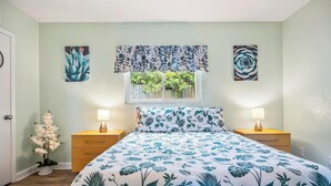 Une chambre à coucher comprenant un lit, deux tables de chevet avec des lampes, une fenêtre donnant sur un paysage verdoyant et deux œuvres d’art murales.