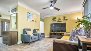 Un salon comprenant un canapé, un téléviseur, un ventilateur de plafond et un coin cuisine avec un réfrigérateur.