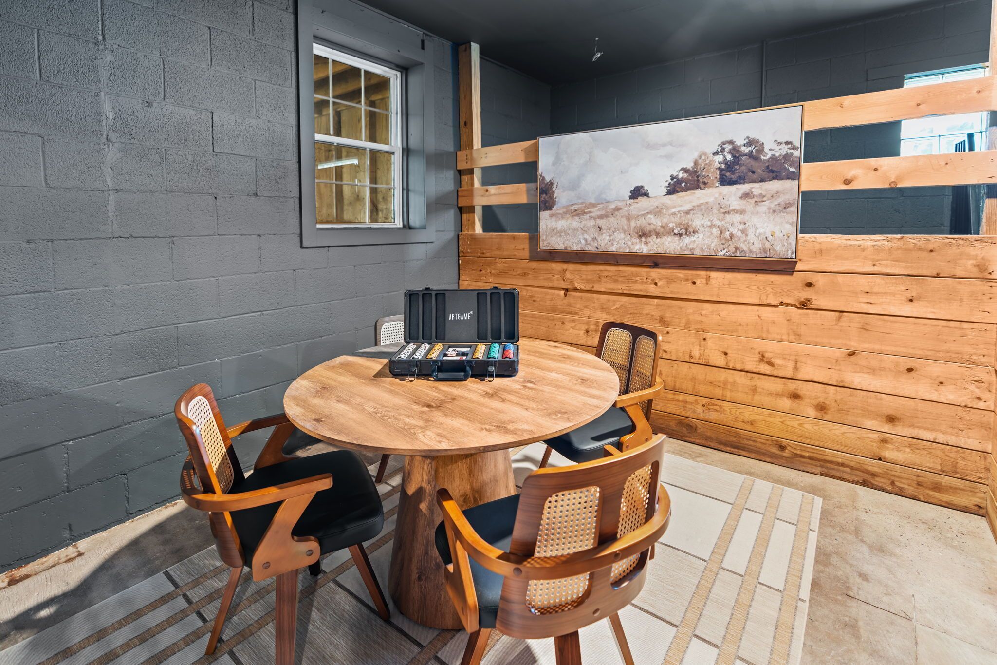 A round wooden table with four chairs, a black suitcase, and a framed picture on the wall.