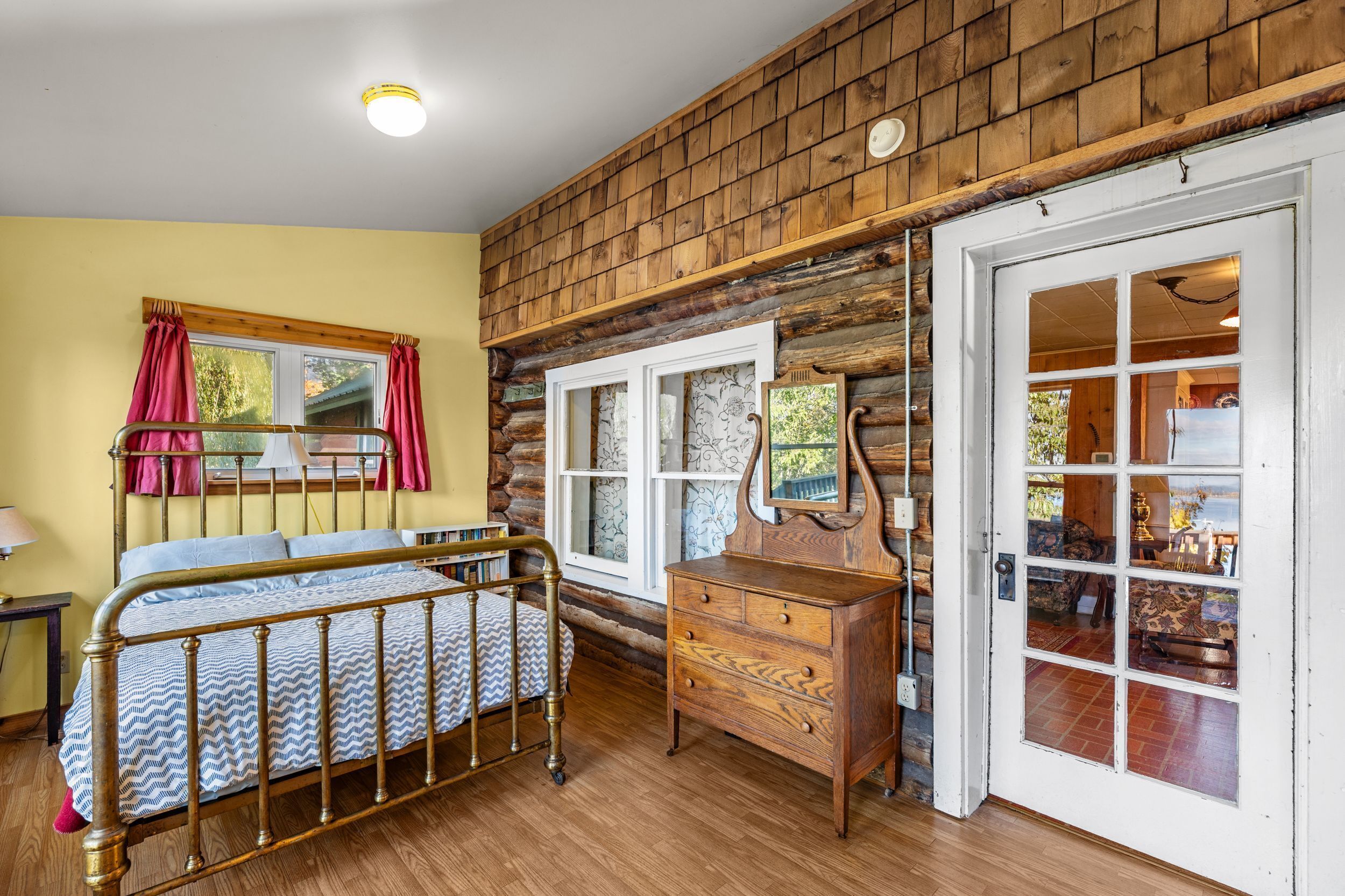 A room with a wooden log wall, a bed with a metal frame, a wooden dresser, and a door leading to another room.