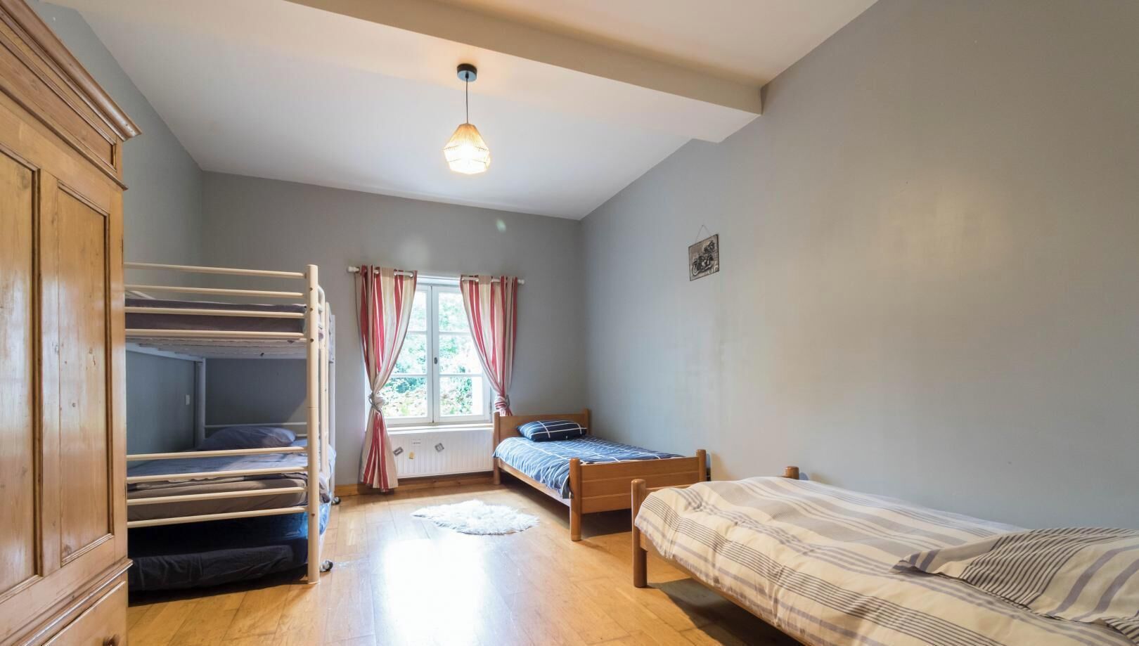 Une chambre de dortoir avec des lits superposés, un lit simple, une fenêtre avec des rideaux, une armoire en bois et un luminaire suspendu.
