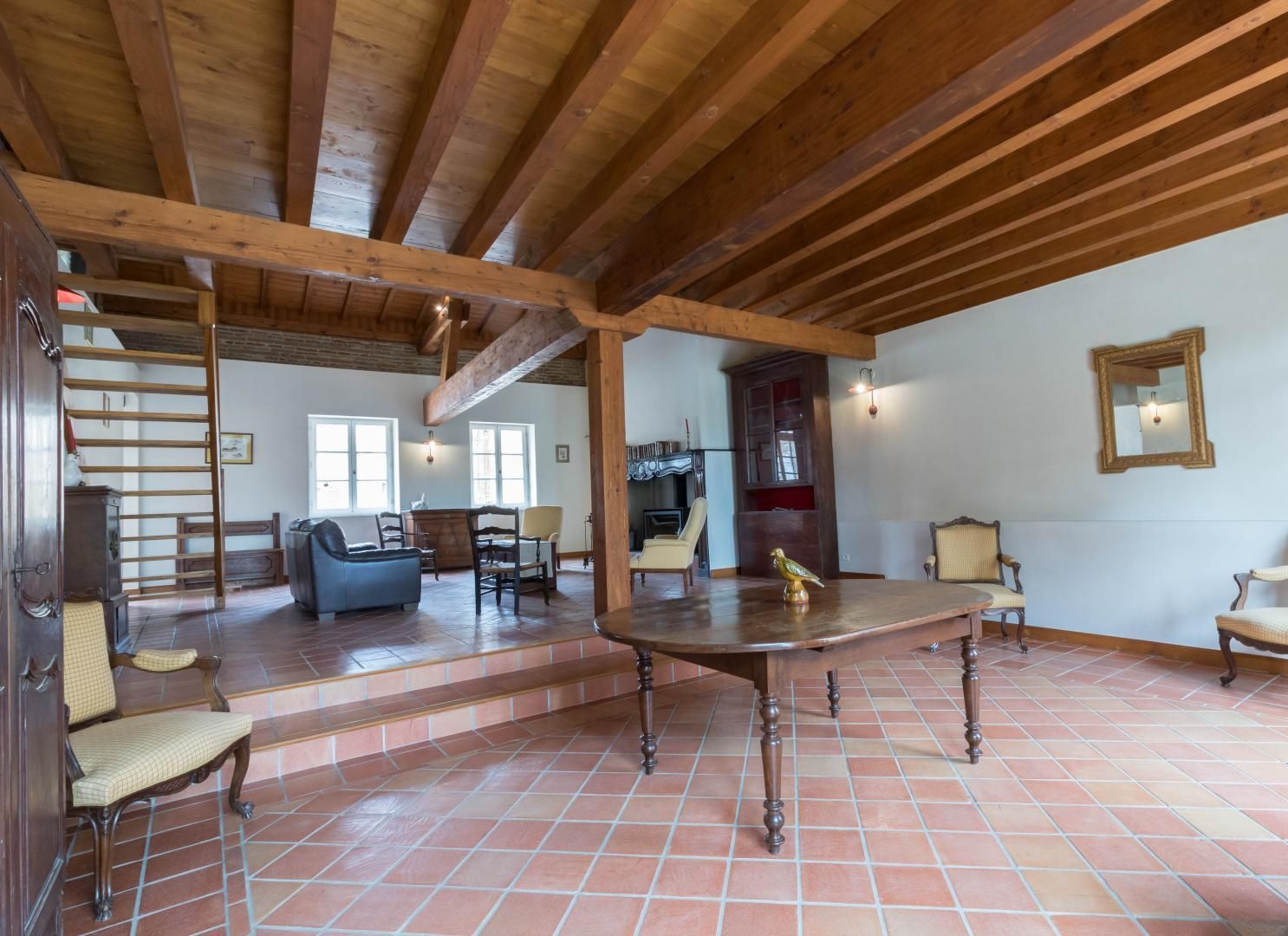 Uma sala de estar ampla, com teto alto e vigas de madeira aparentes, lareira e mesa de jantar.