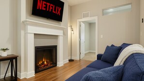 A living room with a fireplace, a blue sofa, and a TV mounted above the fireplace displaying "NETFLIX".