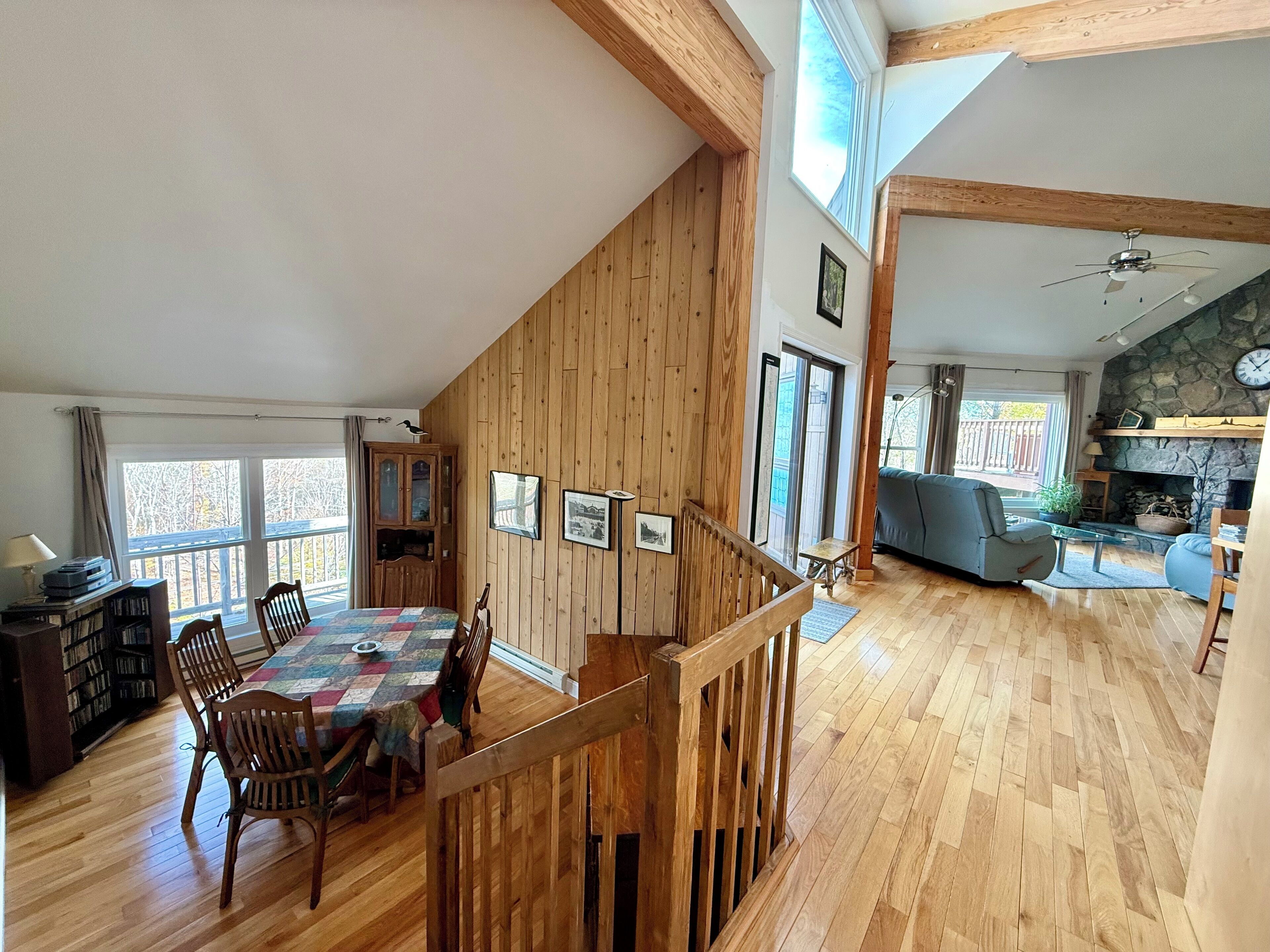 A spacious living area with a wooden floor, a dining table with chairs, a staircase, and a stone fireplace.
