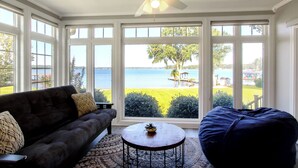 Una habitación con un sofá de color oscuro, una mesa redonda y un sillón de saco. Amplias ventanas ofrecen vistas a un paisaje verde y una masa de agua.