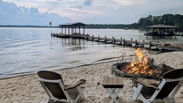 Una playa con dos sillas Adirondack, un brasero y un muelle de madera que se extiende sobre aguas tranquilas.