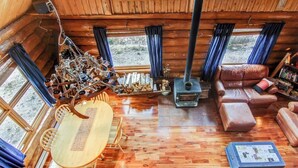 A cozy cabin interior with a wooden dining table, a sofa, a fireplace, and a bookshelf.