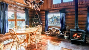 A wooden cabin interior with a dining table, chairs, a chandelier, a fireplace, and large windows.