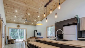 A modern kitchen with wooden ceiling, pendant lights, and a large window overlooking a forest.