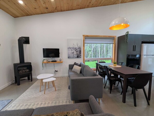 A modern living room with a grey sofa, a dining table, and a TV mounted on the wall. - Hauska | Tavata Chalets | Rest and Nature (Saint-David-de-Falardeau)