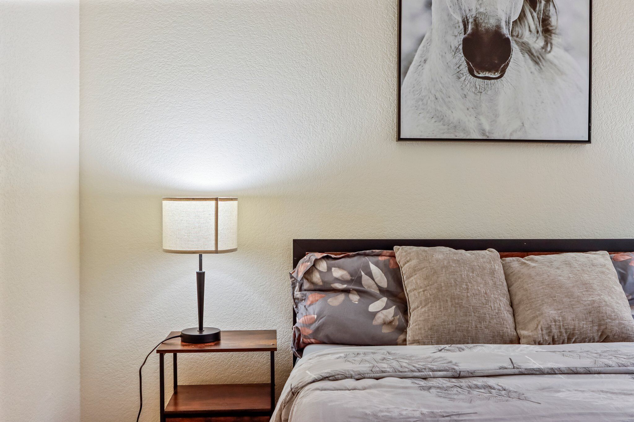 A neatly made bed with a floral-patterned duvet, a bedside lamp, and a framed picture on the wall.