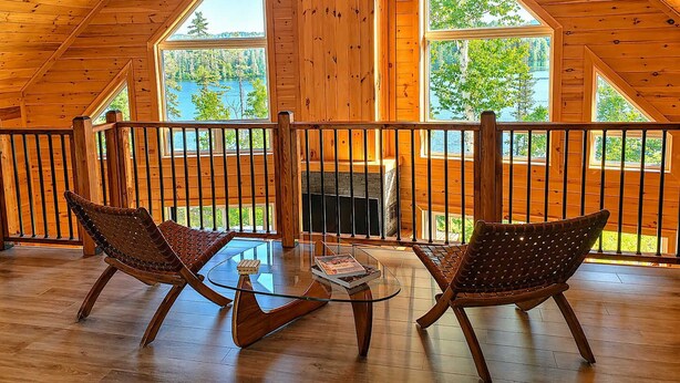 A wooden cabin with a balcony, two wicker chairs, a glass coffee table, and a fireplace.