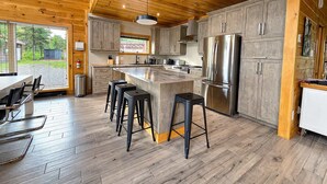 A modern kitchen with wooden ceiling beams, a central island with stools, stainless steel appliances, and a large window.