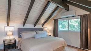 A bedroom with a large bed, two bedside tables with lamps, a window with curtains, and an air conditioning unit.