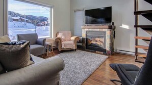 A cozy living room with a fireplace, a large window offering a view of a snowy landscape, and a flat-screen TV mounted above the fireplace.