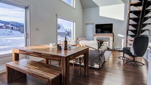 A spacious living room with a wooden dining table, a fireplace, a TV, and a large window offering a view of a snowy landscape.